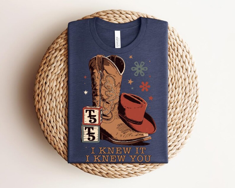 May include: A navy blue t-shirt featuring a graphic of a brown cowboy boot, a red cowboy hat, and two stacked alphabet blocks with the letter T. The design includes small stars and flower shapes, with the text I KNEW IT I KNEW YOU printed below.