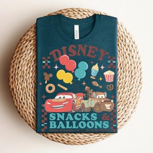 May include: Teal t-shirt with a graphic featuring the word "Disney" above colorful balloons, snacks, and cartoon cars. The text "Snacks & Balloons" is below the cars. The shirt is displayed on a woven, circular surface.
