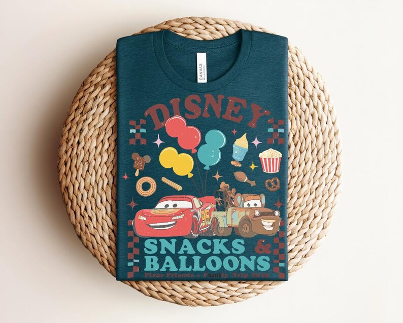 May include: Teal t-shirt with a graphic featuring the word "Disney" above colorful balloons, snacks, and cartoon cars. The text "Snacks & Balloons" is below the cars. The shirt is displayed on a woven, circular surface.