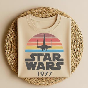 May include: Beige t-shirt featuring a retro design with a colorful sunset, a silhouette of a spaceship, and the words "STAR WARS 1977" in a distressed font. The shirt is folded on a woven mat.
