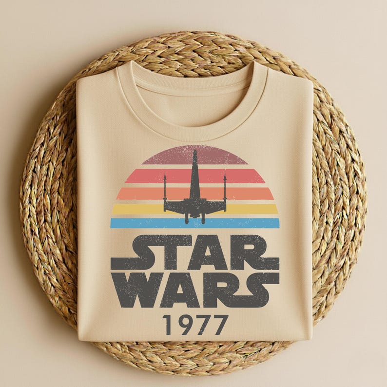 May include: Beige t-shirt featuring a retro design with a colorful sunset, a silhouette of a spaceship, and the words "STAR WARS 1977" in a distressed font. The shirt is folded on a woven mat.