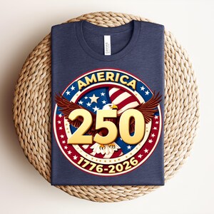 May include: A navy blue t-shirt with a circular graphic. The graphic includes the word "AMERICA", stars, stripes, and an eagle. The number "250" is in gold, with the years "1776-2026". The shirt is folded on a woven surface.