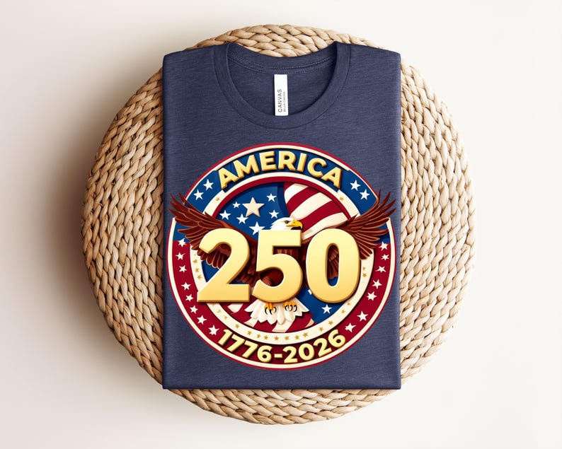May include: A navy blue t-shirt with a circular graphic. The graphic includes the word "AMERICA", stars, stripes, and an eagle. The number "250" is in gold, with the years "1776-2026". The shirt is folded on a woven surface.