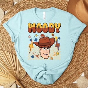 Puede incluir: Camiseta azul claro con la palabra "WOODY" en fuente retro marr&oacute;n y amarilla. La camiseta presenta una ilustraci&oacute;n de dibujos animados de un vaquero con sombrero marr&oacute;n, estrellas y rayos.