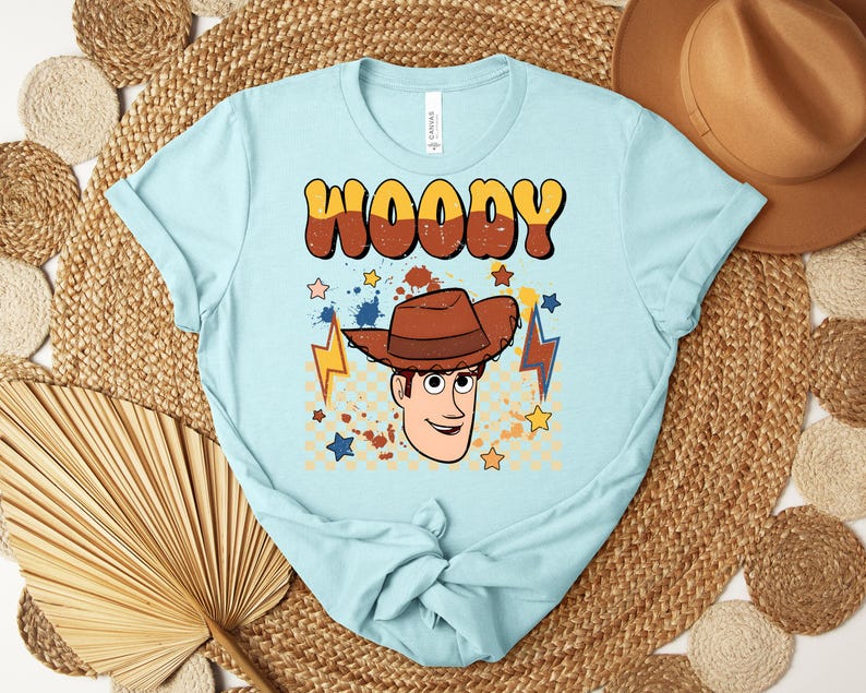 Puede incluir: Camiseta azul claro con la palabra "WOODY" en fuente retro marr&oacute;n y amarilla. La camiseta presenta una ilustraci&oacute;n de dibujos animados de un vaquero con sombrero marr&oacute;n, estrellas y rayos.