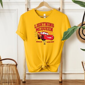 May include: Yellow t-shirt with a red cartoon race car and the text "Lightning McQueen" in red and black. The car is holding a trophy and the text "Piston Cup Racing Champion" is below the car.