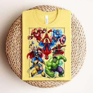 May include: A folded yellow t-shirt with a vibrant graphic of Marvel superheroes such as Iron Man, Captain America, and Spider-Man. The design is framed within a rectangle. The shirt rests on a circular woven mat.