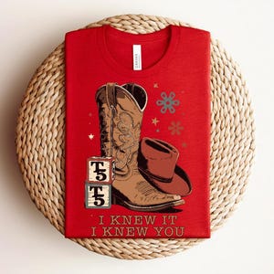 May include: A red t-shirt featuring a graphic of a brown cowboy boot, a brown cowboy hat, and two stacked alphabet blocks with the letter T and the number 5. The design includes small star and flower icons and the text I KNEW IT I KNEW YOU.