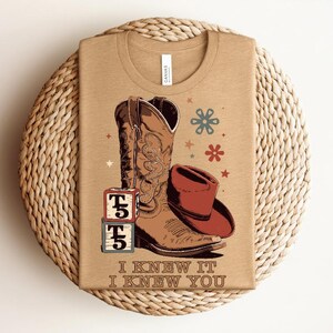 May include: A tan t-shirt featuring a graphic of a brown cowboy boot, a red cowboy hat, and two stacked alphabet blocks with the letter T and the number 5. The design includes small star and flower icons and the text I KNEW IT I KNEW YOU.