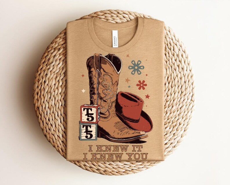 May include: A tan t-shirt featuring a graphic of a brown cowboy boot, a red cowboy hat, and two stacked alphabet blocks with the letter T and the number 5. The design includes small star and flower icons and the text I KNEW IT I KNEW YOU.