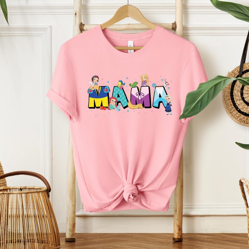 K&ouml;nnte beinhalten: Rosafarbenes T-Shirt mit einem Aufdruck von Disney-Prinzessinnen, die das Wort "MAMA" buchstabieren.