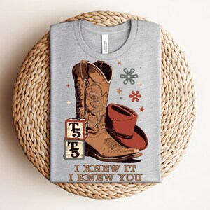 May include: A heather gray t-shirt featuring a graphic of a brown cowboy boot, a red cowboy hat, two stacked alphabet blocks with the letter T, and small floral and star icons. The text "I KNEW IT I KNEW YOU" is printed in brown at the bottom.