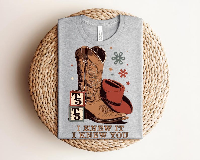 May include: A heather gray t-shirt featuring a graphic of a brown cowboy boot, a red cowboy hat, two stacked alphabet blocks with the letter T, and small floral and star icons. The text "I KNEW IT I KNEW YOU" is printed in brown at the bottom.