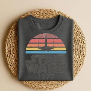 May include: Dark gray t-shirt featuring a retro design with a colorful sunset, a silhouette of a spaceship, and the text "STAR WARS 1977". The shirt is folded on a woven mat.