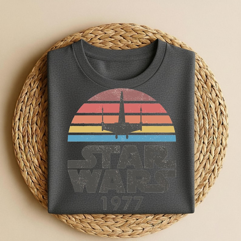 May include: Dark gray t-shirt featuring a retro design with a colorful sunset, a silhouette of a spaceship, and the text "STAR WARS 1977". The shirt is folded on a woven mat.