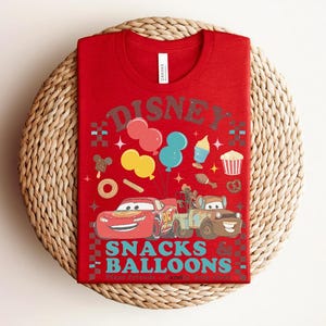 May include: Red t-shirt featuring a graphic of Lightning McQueen and Mater from the movie Cars, along with balloons and snacks. The shirt has the word "Disney" above the graphic and "Snacks Balloons" below. The shirt is folded on a woven mat.