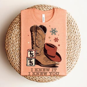 May include: A peach-colored t-shirt featuring a graphic of a brown cowboy boot, a brown cowboy hat, and two stacked blocks with the letter T and the number 5. The text "I KNEW IT I KNEW YOU" is printed below the graphic with small flower icons.