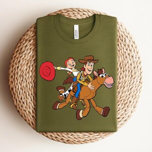 May include: Olive green t-shirt featuring a graphic of Woody, Jessie, and a horse from Toy Story. Woody is wearing a cowboy hat and vest, while Jessie holds a red hat. The shirt is displayed on a woven surface.