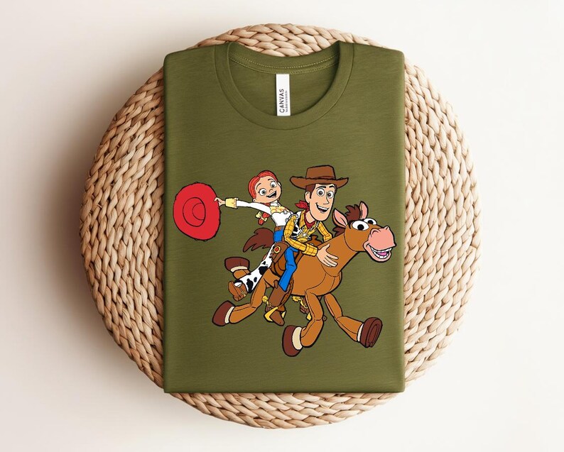 May include: Olive green t-shirt featuring a graphic of Woody, Jessie, and a horse from Toy Story. Woody is wearing a cowboy hat and vest, while Jessie holds a red hat. The shirt is displayed on a woven surface.