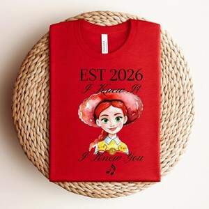 Pode incluir: Uma t-shirt vermelha com o gr&aacute;fico de uma personagem de desenho animado com cabelo ruivo e um chap&eacute;u de aba larga. O texto diz EST 2026, I Knew It e I Knew You, acompanhado por uma pequena nota musical. A t-shirt tem gola redonda e uma etiqueta.