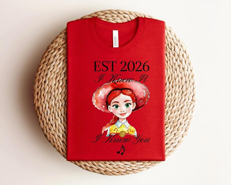 Pode incluir: Uma t-shirt vermelha com o gr&aacute;fico de uma personagem de desenho animado com cabelo ruivo e um chap&eacute;u de aba larga. O texto diz EST 2026, I Knew It e I Knew You, acompanhado por uma pequena nota musical. A t-shirt tem gola redonda e uma etiqueta.