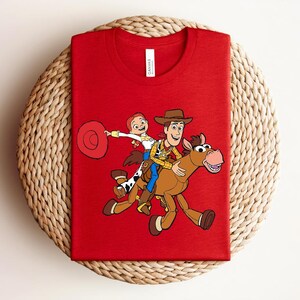 May include: A red t-shirt featuring a graphic of Woody, Jessie, and a horse from the Toy Story movie. The characters are in full color, with Woody wearing a cowboy hat and Jessie holding a lasso. The shirt is made of a soft material.