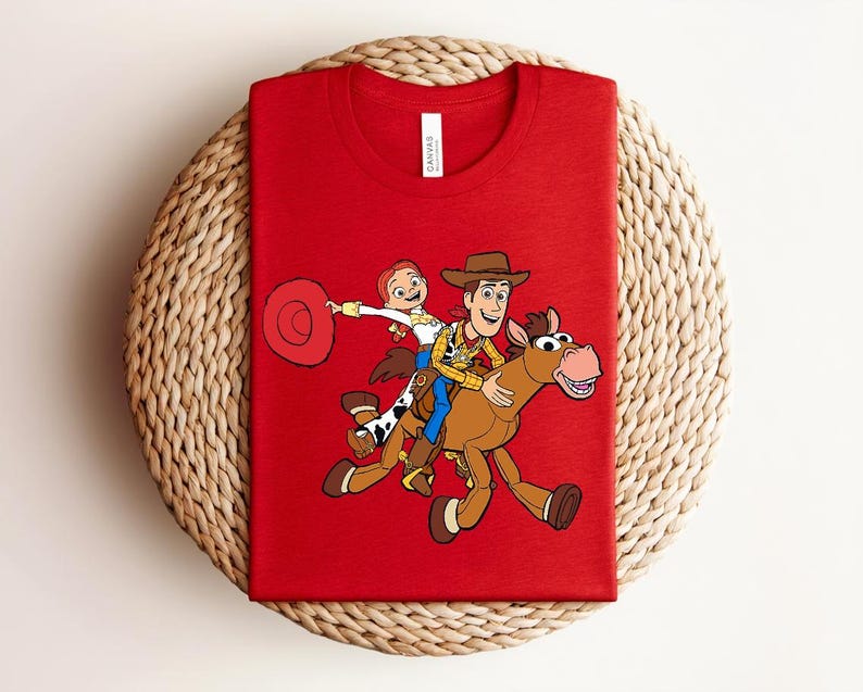 May include: A red t-shirt featuring a graphic of Woody, Jessie, and a horse from the Toy Story movie. The characters are in full color, with Woody wearing a cowboy hat and Jessie holding a lasso. The shirt is made of a soft material.