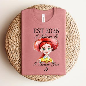 Pode incluir: Uma t-shirt rosa seco com o gr&aacute;fico de uma personagem ruiva a usar um chap&eacute;u vermelho e uma camisa amarela. O texto diz EST 2026, I Knew It e I Knew You, acompanhado por uma pequena nota musical preta sob a ilustra&ccedil;&atilde;o.