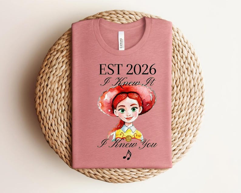 Pode incluir: Uma t-shirt rosa seco com o gr&aacute;fico de uma personagem ruiva a usar um chap&eacute;u vermelho e uma camisa amarela. O texto diz EST 2026, I Knew It e I Knew You, acompanhado por uma pequena nota musical preta sob a ilustra&ccedil;&atilde;o.