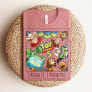 K&ouml;nnte beinhalten: Ein altrosa T-Shirt mit einer Grafik der Toy Story-Figuren, darunter Woody, Buzz Lightyear und Slinky Dog. Das Design enth&auml;lt den Text Toy Story 5 und den Schriftzug I KNEW IT, I KNEW YOU in schwarzen Buchstaben unter den Charakteren.