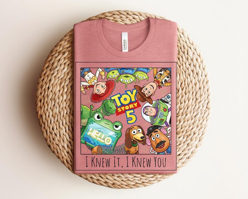 K&ouml;nnte beinhalten: Ein altrosa T-Shirt mit einer Grafik der Toy Story-Figuren, darunter Woody, Buzz Lightyear und Slinky Dog. Das Design enth&auml;lt den Text Toy Story 5 und den Schriftzug I KNEW IT, I KNEW YOU in schwarzen Buchstaben unter den Charakteren.