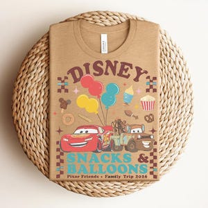 May include: A tan t-shirt featuring a graphic design with the text "Disney Snacks & Balloons". The design includes cartoon cars, balloons, and various food icons. The shirt is laid on a woven, circular placemat.