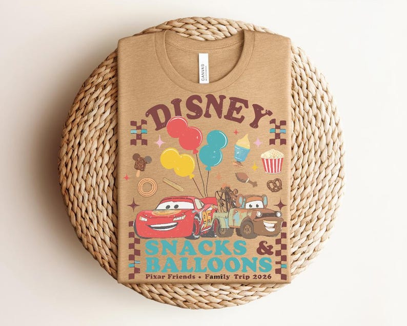 May include: A tan t-shirt featuring a graphic design with the text "Disney Snacks & Balloons". The design includes cartoon cars, balloons, and various food icons. The shirt is laid on a woven, circular placemat.