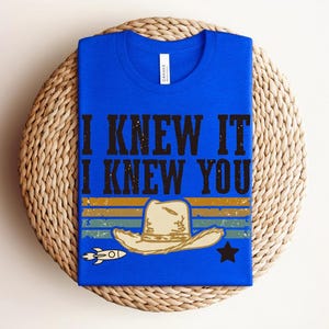 May include: A bright blue t-shirt featuring the black text I KNEW IT I KNEW YOU above a graphic of a beige cowboy hat centered over horizontal stripes in yellow, teal, and blue, with a small white rocket and a black star on the sides.