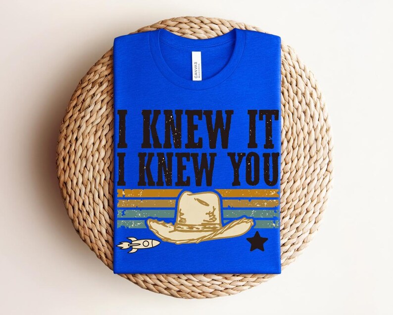May include: A bright blue t-shirt featuring the black text I KNEW IT I KNEW YOU above a graphic of a beige cowboy hat centered over horizontal stripes in yellow, teal, and blue, with a small white rocket and a black star on the sides.
