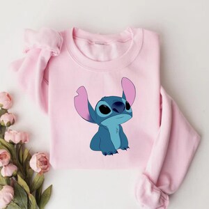 K&ouml;nnte beinhalten: Ein hellrosa Sweatshirt mit einer Cartoon-Illustration von Stitch aus Lilo & Stitch. Die blaue und rosa Figur befindet sich mittig auf der Vorderseite des Sweatshirts. Die &Auml;rmel sind auf beiden Seiten des Sweatshirts sichtbar.