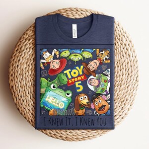 K&ouml;nnte beinhalten: Ein marineblaues T-Shirt mit einem Grafikdruck von Toy Story-Figuren, darunter Woody, Buzz Lightyear, Jessie, Rex, Slinky Dog, Mr. Potato Head und die Aliens. Der Text lautet "TOY STORY 5" und "I KNEW IT, I KNEW YOU" mit einem gr&uuml;nen Tablet.