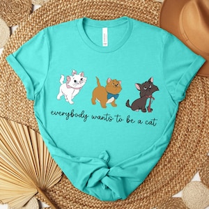 May include: Teal t-shirt featuring three cartoon cats with the text "everybody wants to be a cat". The cats are white, brown, and black, each with unique accessories. The shirt is a crew neck and has a relaxed fit.