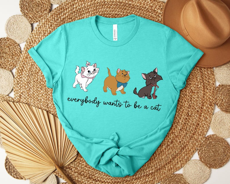 May include: Teal t-shirt featuring three cartoon cats with the text "everybody wants to be a cat". The cats are white, brown, and black, each with unique accessories. The shirt is a crew neck and has a relaxed fit.