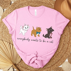 May include: Pink t-shirt featuring three cartoon cats in white, brown, and black. The text "everybody wants to be a cat" is printed below the cats. The shirt is made of a soft material.