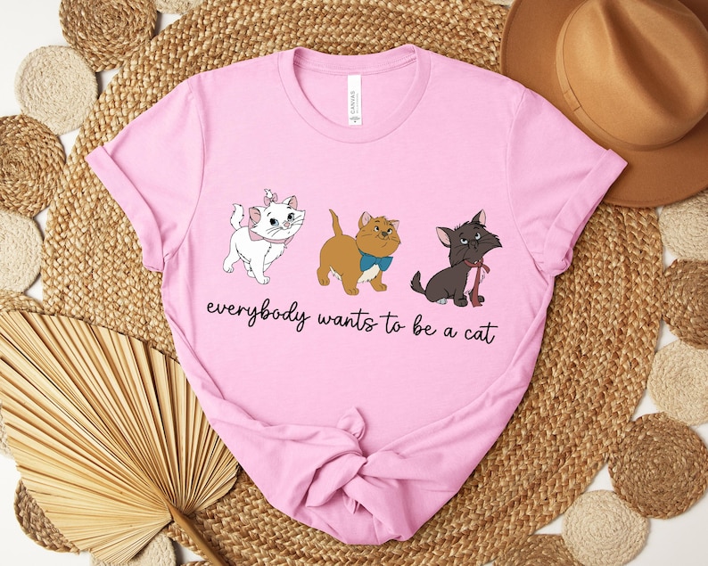 May include: Pink t-shirt featuring three cartoon cats in white, brown, and black. The text "everybody wants to be a cat" is printed below the cats. The shirt is made of a soft material.