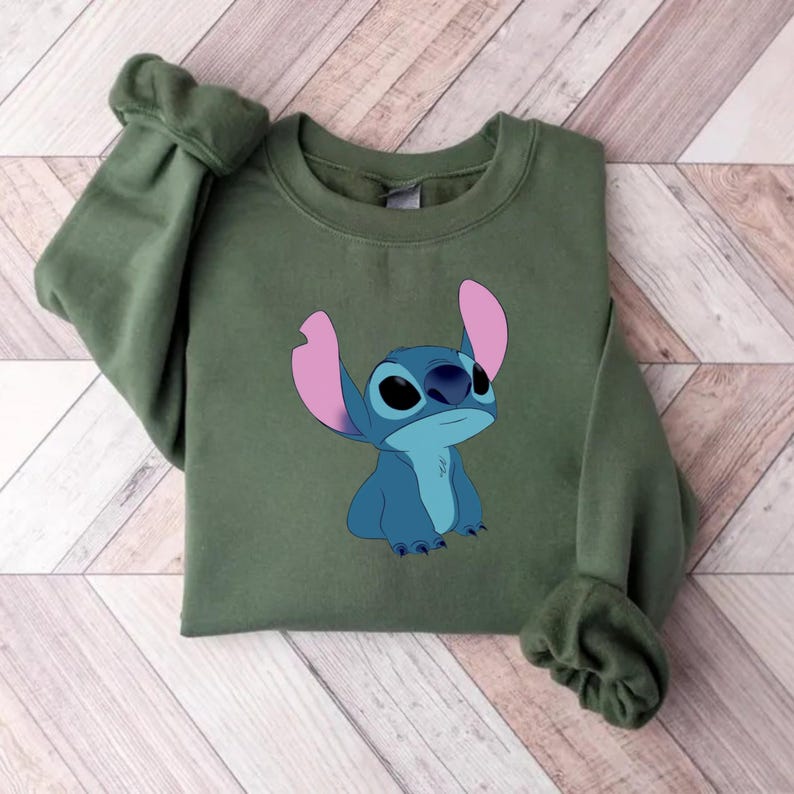 K&ouml;nnte beinhalten: Ein olivgr&uuml;nes Sweatshirt mit einem Cartoon-Bild von Stitch aus Lilo & Stitch. Die Figur ist blau mit gro&szlig;en rosa Ohren. Das Sweatshirt ist auf einer Holzoberfl&auml;che gefaltet.