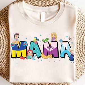 K&ouml;nnte beinhalten: Ein wei&szlig;es T-Shirt mit einer bunten Grafik von Disney-Prinzessinnen, die das Wort "MAMA" buchstabieren. Die Prinzessinnen sind Schneewittchen, Arielle, Rapunzel, Elsa und Anna.