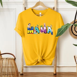 K&ouml;nnte beinhalten: Gelbes T-Shirt mit einem Aufdruck des Wortes "MAMA", das aus Disney-Prinzessinnen besteht. Jeder Buchstabe hat eine andere Farbe und zeigt eine andere Prinzessin.