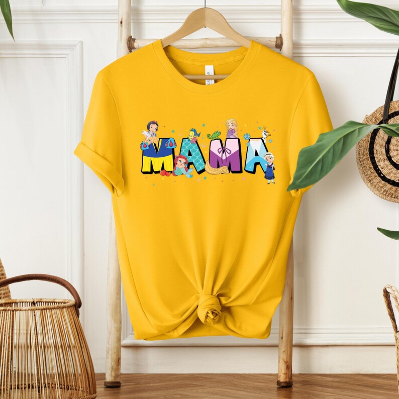 K&ouml;nnte beinhalten: Gelbes T-Shirt mit einem Aufdruck des Wortes "MAMA", das aus Disney-Prinzessinnen besteht. Jeder Buchstabe hat eine andere Farbe und zeigt eine andere Prinzessin.