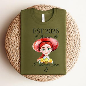 Pode incluir: Uma t-shirt verde-oliva com uma ilustra&ccedil;&atilde;o em estilo aguarela de uma personagem ruiva com um chap&eacute;u vermelho. O texto diz EST 2026, I Knew It e I Knew You, acompanhado por um pequeno s&iacute;mbolo de nota musical impresso em tinta preta.