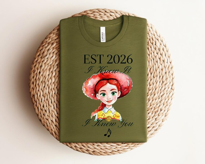 Pode incluir: Uma t-shirt verde-oliva com uma ilustra&ccedil;&atilde;o em estilo aguarela de uma personagem ruiva com um chap&eacute;u vermelho. O texto diz EST 2026, I Knew It e I Knew You, acompanhado por um pequeno s&iacute;mbolo de nota musical impresso em tinta preta.
