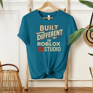 May include: Teal t-shirt with the text "BUILT DIFFERENT in ROBLOX STUDIO" in a retro font. The design includes a lightning bolt and a red square. The shirt is hanging on a wooden hanger.