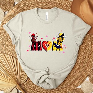 Op de afbeelding: Een lichtbeige t-shirt met een grafisch ontwerp met het woord "MOM" verdeeld over twee superheldenpersonages. De "M" toont Deadpool, de "O" is een hart en de "M" toont Wolverine. Roze hartjes zijn verspreid rond het ontwerp.
