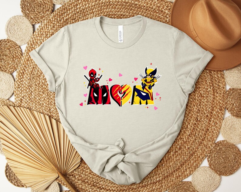 Op de afbeelding: Een lichtbeige t-shirt met een grafisch ontwerp met het woord "MOM" verdeeld over twee superheldenpersonages. De "M" toont Deadpool, de "O" is een hart en de "M" toont Wolverine. Roze hartjes zijn verspreid rond het ontwerp.
